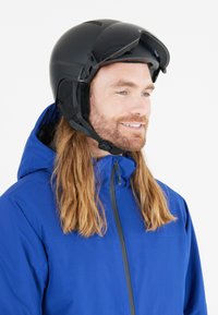 Sort skihjelm med visir, strømlinet design, justerbar hagekomfort, båret med en blå jakke med høj krave og lynlås.