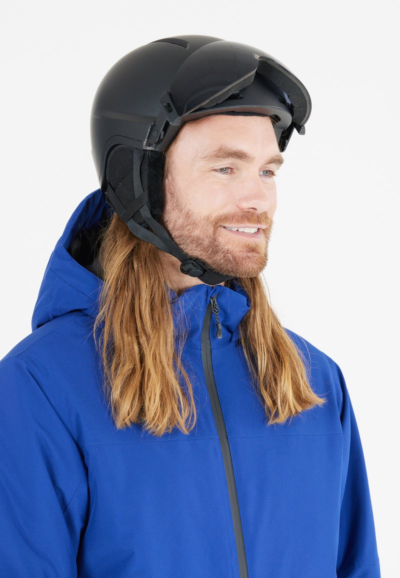 Sort skihjelm med visir, strømlinet design, justerbar hagekomfort, båret med en blå jakke med høj krave og lynlås.