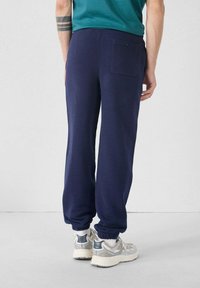 Marineblauwe joggingbroek van zacht materiaal met elastische boorden, met een achterzak met een knoopdetail. Draagt bij lichtgekleurde sneakers.