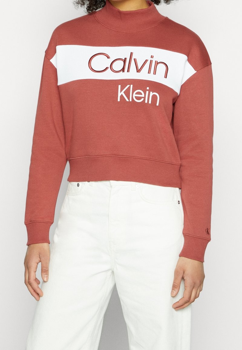 Sweat-shirt Calvin Klein couleur rouille avec bande blanche, col montant, poignets élastiques et coupe courte. Présente un logo aux couleurs contrastantes.