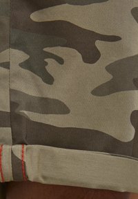 Shorts camouflage avec un mélange de tons vert olive, beige et marron ; présentent un bord roulé avec un détail de couture rouge.