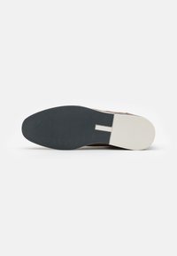 Zapato de cuero marrón con suela de goma negra texturizada y detalle en el talón blanco. El diseño es plano con una forma limpia y sencilla.