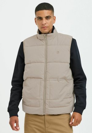 Beige Steppweste mit hohem Kragen, frontalem Reißverschluss, zwei Taschen und genähten Details. Getragen über einem schwarzen Langarmshirt.