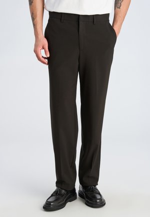 Mann trägt schwarze maßgeschneiderte Hose und schwarze Leder-Troddel-Loafer, steht mit einer Hand in der Tasche vor einfachem Hintergrund.