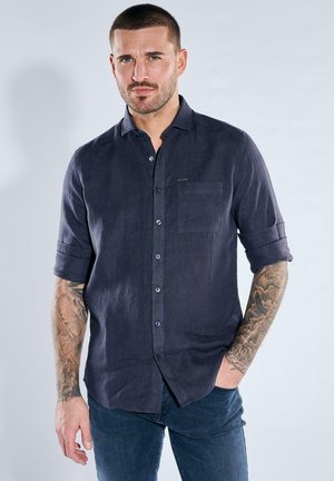 Mann mit kurzem Haar und Tätowierungen trägt ein dunkelblaues Hemd mit aufgerollten Ärmeln und dunkle Jeans, steht vor einem hellen Hintergrund.