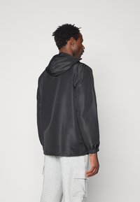 Jacket preto em nylon com capuz, punhos elásticos e corte solto. Tecido texturizado, design simples sem padrões ou detalhes visíveis.