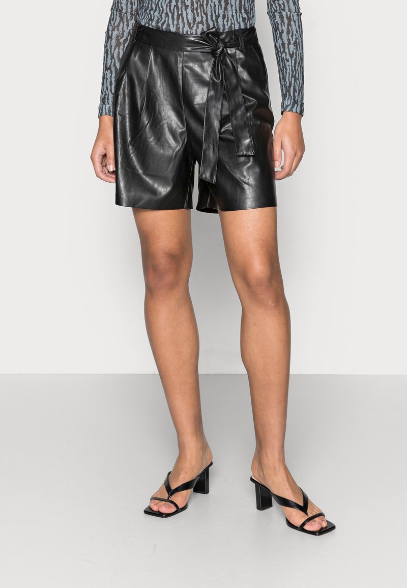 Femme portant un short en cuir noir à taille nouée, des sandales à talons noires et un haut à manches longues à motifs, debout devant un fond uni.