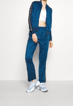 Personne portant une veste et un pantalon de survêtement bleus avec des bandes blanches au logo, un crop top blanc et des baskets blanches, debout avec une main dans la poche.