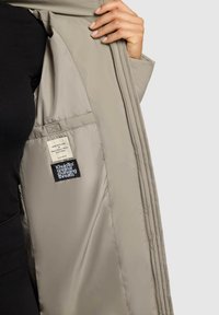 Giacca a vento beige chiaro con una texture liscia, che mostra un'etichetta interna e dettagli di cucitura, tenuta aperta per rivelare la fodera e le tasche.