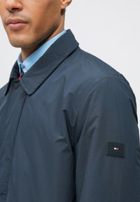 Muž v tmavomodrom saku s preloženým golierom a svetlomodrou košeľou pod ním, na rukáve ukazujúc malý logotyp.