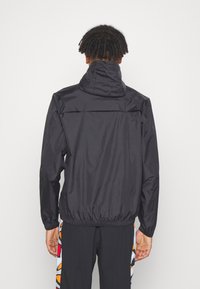 Schwarze, leichte Jacke mit hohem Kragen. Gummizüge an den Ärmeln und dem Saum. Flüssige, glänzende Textur. Getragen mit schwarzen Hosen mit bunten Akzenten.