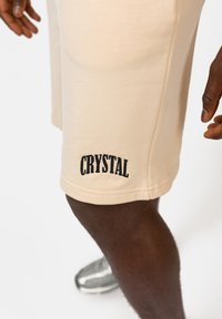 Beige katoenen shorts met zwarte "CRYSTAL" borduursel op de zoom. De stof is soepel en heeft een relaxte pasvorm.