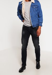 Jeansjacka med en beige shearlingkrage, blåtvätt, framfickor; grå T-shirt, mörka jeans och svarta ankelskor fulländar outfiten.