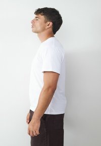 Weißen Baumwoll-T-Shirt mit kurzen Ärmeln, Rundhalsausschnitt und lockerer Passform. Kombiniert mit dunkelbraunen Hosen.