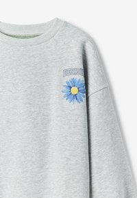 Felpa grigia chiara con colletto a costine, caratterizzata da una grafica di margherita blu e gialla e parte del testo "DESIGU" sul lato superiore sinistro del petto.