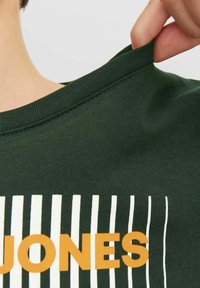 Grøn bomulds T-shirt med ribbet krave, der har lodrette hvide striber og ordet "JONES" i orange på fronten.