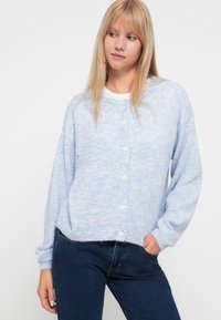 Lys blå, blød strikcardigan med lange ærmer, rund halsudskæring og fem hvide knapper. Båret over en hvid skjorte, parret med mørke jeans.