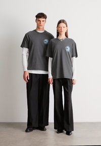 Γκρι υπερμεγέθη μπλουζάκια με στρογγυλή λαιμόκοψη και τυπωμένο λογότυπο, συνδυασμένα με σκούρα φαρδιά παντελόνια, φορεμένα από δύο μοντέλα μπροστά σε απλό φόντο.