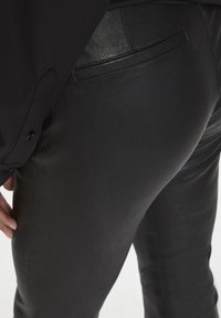 Schwarze Lederhosen mit glatter Textur, mit einer Gesäßtasche und einer figurbetonten Passform. Beinhaltet eine partielle Sicht auf einen Ärmel eines schwarzen Shirts.