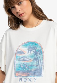 Bílý bavlněný tričko s vintage stylem grafiky, na které jsou zobrazeny palm tree, surfaři a mraky v modrých a oranžových odstínech. Text "ROXY" v dolní části.