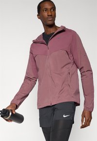 Ελαφρύ, μαρόν μπουφάν με φερμουάρ και κουκούλα, με υφή από δίχτυ και λογότυπο. Συνδυάζεται με μαύρα σορτς και κρατάει ένα σκουρόχρωμο παγούρι νερού.