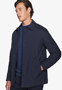 Marineblå letvægtsjakke med krave, knaplukninger, to sidelommer og lige skæring. Bæres over en blå sweater.