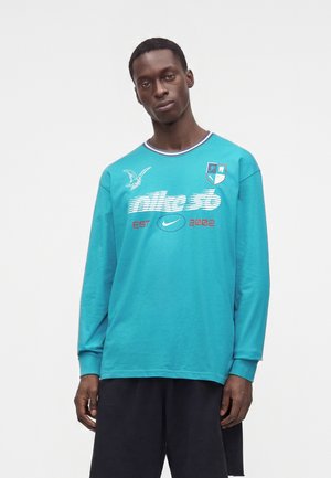 Homme portant un t-shirt à manches longues turquoise de Nike avec logo, graphique d'aigle, et un short noir, debout devant un fond blanc uni.