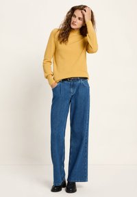 Frau mit lockigem Haar, die einen senfgelben Rollkragenpullover, weite blaue Jeans, einen schwarzen Gürtel und schwarze Schuhe trägt, steht vor einem cremefarbenen Hintergrund.