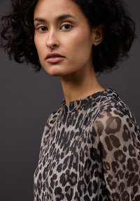 Halenka s leopardím vzorem, vysokým límcem, průhlednými rukávy a texturovaným materiálem. Barevná paleta zahrnuje béžové, hnědé a černé skvrny.