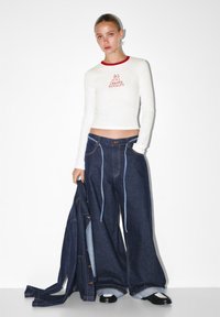 Valkoinen pitkähihaista paita, jossa on punainen kaulus ja brodeerattu graafinen kuvio, yhdistettynä ylisuuria tummansinisiä farkkuhousuja, joissa on vaaleansiniset stitchingit ja lahkeet.