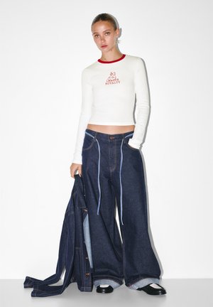 Biely dlhý rukáv top s červeným golierom a vyšitým grafickým motívom, v kombinácii s nadrozmernými tmavomodrými džínsami so svetlomodrým prešívaním a manžetami.