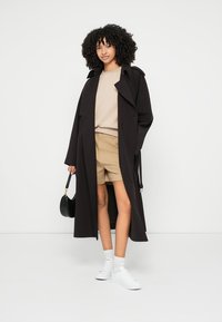 Črni dolg trench plašč, krem pulover, bež kratke hlače, beli supergi in mala črna torbica. Gladek material in sproščen kroj.