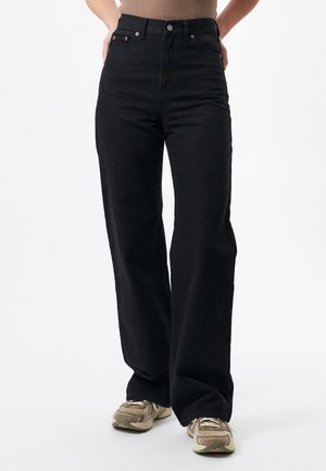 Black wide-leg jeans made from denim, featuring a high waist, copper hardware, and dual front pockets, paired with tan sneakers.