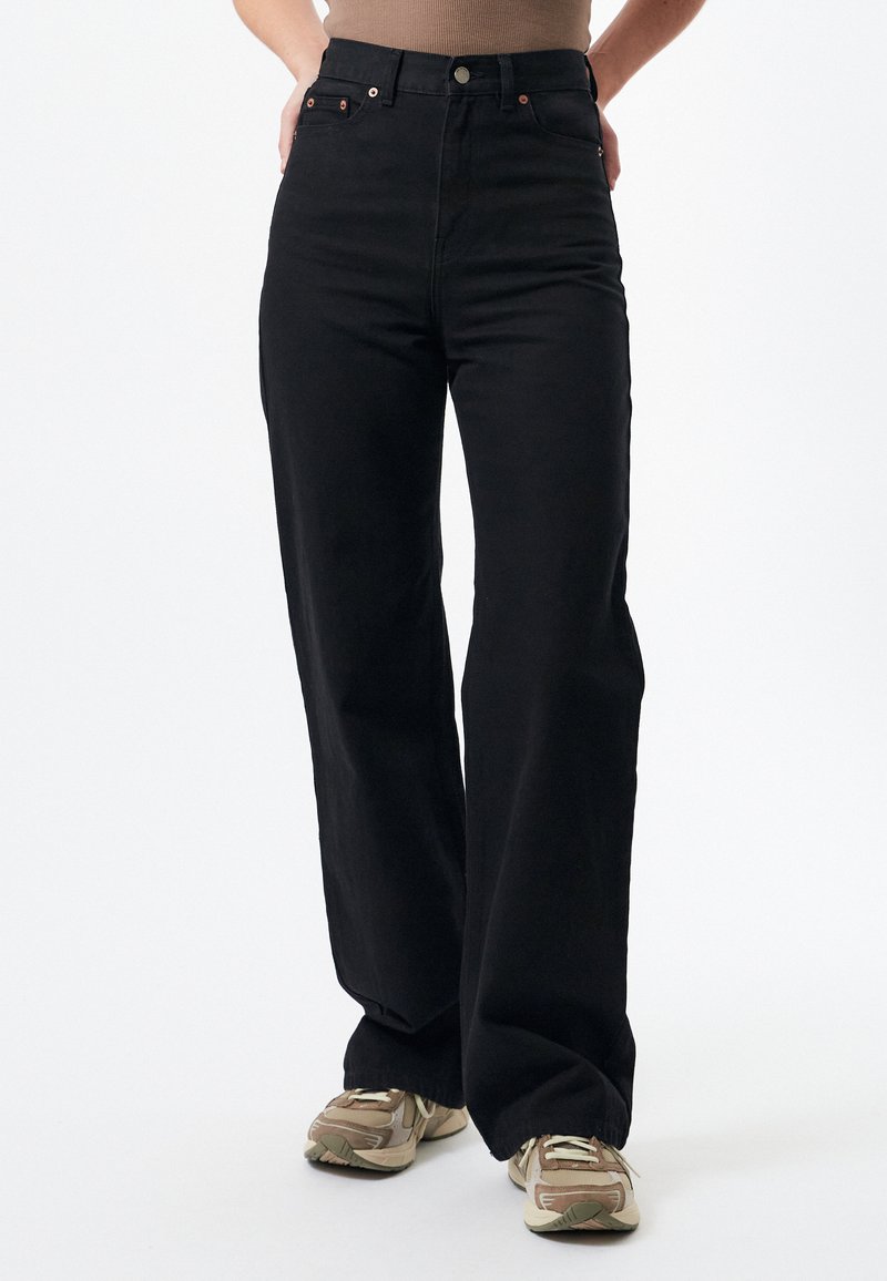 Black wide-leg jeans made from denim, featuring a high waist, copper hardware, and dual front pockets, paired with tan sneakers.