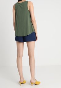 Woman standing facing away, wearing sleeveless olive green top, navy blue shorts, and yellow heeled mule shoes against plain background.