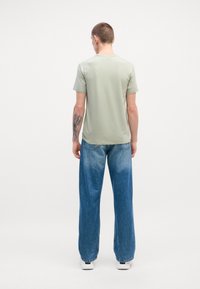 T-shirt vert clair à manches courtes, texture lisse, associé à un jean en denim bleu ample avec un dégradé et des baskets blanches.