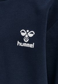 Jūras spēku krāsas džemperis ar mīkstu tekstūru, uz kura priekšpuses centrā atrodas balta Hummel logo. Vienkāršs dizains ar apaļu kakla izgriezumu.