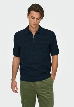 Uomo che indossa una polo blu navy a maniche corte con texture e zip, abbinata a pantaloni verdi di velluto a coste, in piedi con una mano in tasca.