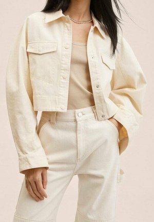 Femme portant une veste courte crème avec des boutons-pression, un débardeur côtelé beige et un pantalon crème taille haute, la main reposant dans la poche.