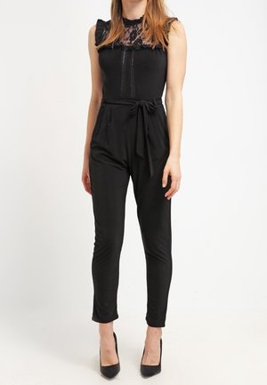 Kobieta ubrana w bezrękawnikową czarną koronkową bluzkę, wysokie czarne spodnie związywane w talii i czarne buty na obcasie.