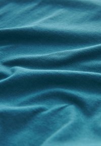 Weicher türkisfarbener Stoff mit sanften Falten und Knicken, der Textur und glatte Oberfläche bei gleichmäßiger Beleuchtung zeigt.