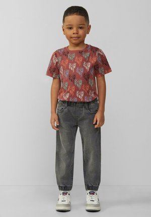 Rood en groen palmgepatroniseerd kortärmige shirt gecombineerd met grijze taps toelopende joggers en witte sneakers met zwarte accenten.