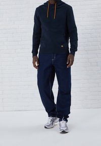 Sudadera azul marino con cordones naranjas y un bolsillo frontal, combinada con jeans de mezclilla azul de corte holgado y zapatillas deportivas plateadas.
