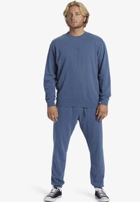 Felpa blu e pantaloni jogger coordinati realizzati in tessuto morbido, con polsini a coste e un piccolo logo sul petto. Abbinati a sneakers nere.