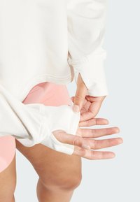 Leichtes, cremefarbenes Langarmoberteil mit Daumenlöchern; glatte Textur; Saum endet über der Taille; kombiniert mit pinken Shorts.