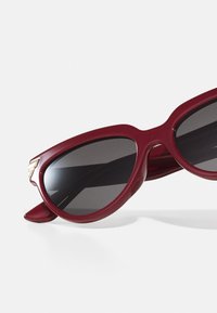 Gafas de sol estilo cat-eye en burdeos con lentes oscuros, marco de plástico suave y detalle en tono dorado en los laterales. Diseño ligero.