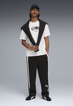 Hombre con gorra negra, camiseta blanca con diseño gráfico, chaqueta negra sobre los hombros, pantalones deportivos negros con rayas blancas y zapatillas negras.