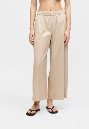 Person iført beige bukser med vide ben og elastisk talje samt brune flade sandaler, stående mod en ensartet lys baggrund.