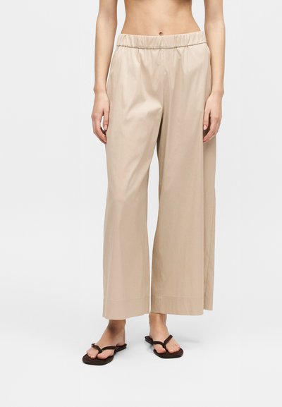 Personne portant un pantalon beige à jambes larges avec une taille élastique et des sandales plates marron, debout devant un fond clair uni.