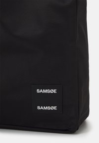 Borsa di stoffa nera con superficie texture. Presenta un'etichetta rettangolare con il nome del marchio "SAM_SØE" in testo bianco. Design semplice e minimale.